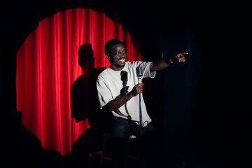 Happy young african american man comic tells stand up monologue on stage with red curtains. Concept humor standup comedian show open mike