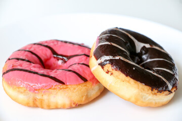 Sweet tooth concept. Two doughnut (donut) rings glazed with pink strawberry and chocolate sugar icing, on white dish. Delicious sugary food but unhealthy with high calories.