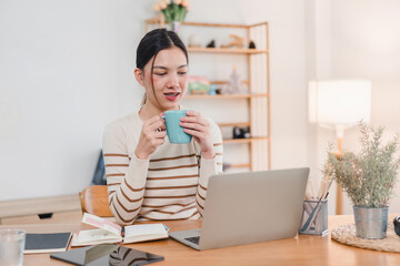 Asian woman sitting comfortably with a cup of hot beverage, smiling and enjoying a cozy moment in home office.