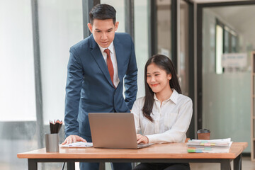 A man and a woman are working together on a laptop. The man is wearing a suit and tie, and the woman is wearing a white shirt. They are both focused on the laptop screen, which is open
