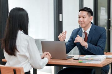 A man and a woman are sitting at a table, talking to each other. The man is wearing a suit and tie, and the woman is wearing a white shirt. They are both looking at a laptop computer
