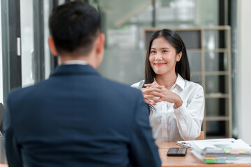 A man and a woman are sitting at a desk. The man is wearing a suit and the woman is wearing a white shirt. They are both smiling and seem to be having a pleasant conversation