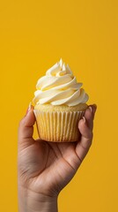 A hand holding a delicious cupcake with creamy frosting against a bright yellow background, showcasing the dessert beautifully.
