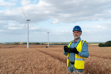 Man in Safety Gear Stands in Wheat Field With Wind Turbines During Sunny Day