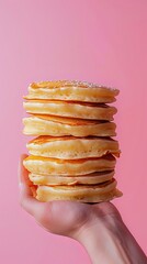 A hand holds a tall stack of fluffy and golden pancakes, topped with a sprinkle of powdered sugar against a vibrant pink background.