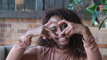 Portrait of a cute African American woman with a beautiful smile, with long curly hair. A woman looks at the camera and shows a heart with her fingers, a symbol of love.
