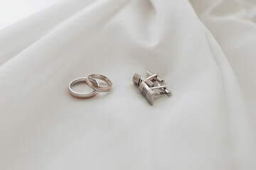 A silver ring and a silver cufflink are on a white cloth. The cufflink has a cross on it