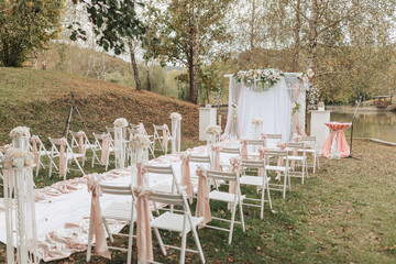A wedding ceremony is taking place in a park with a white tent and a white arch. The chairs are arranged in a long line, and there are many flowers and decorations. Scene is joyful and celebratory