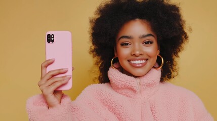 A woman is holding a pink cell phone and smiling. She is wearing a pink jacket and has a gold hoop earring