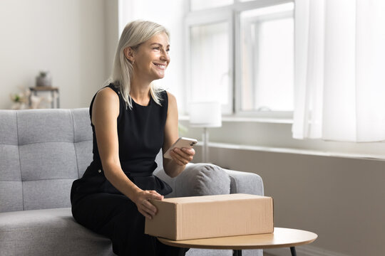 Woman prepare parcel box for send use on-line postal services, sit on sofa hold phone, writes to courier, provide address data, conducting commercial transactions via application send goods to client