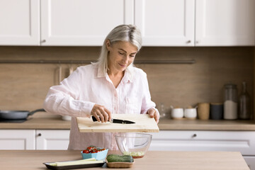Smiling pretty mature woman prepare tasty vegan salad in cozy kitchen, start new day with healthy...