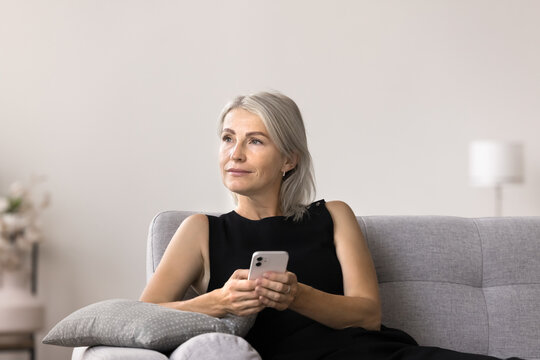 Pensive mature woman sits on sofa holding smartphone looking away thinking, wait for call from friend spend time alone in living room. Modern gadget usage for communication, making order or phonecall
