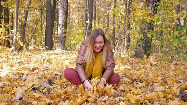Joyful woman in brown jacket with yellow scarf plays among fallen leaves. She tosses yellow leaves into air with excitement, as autumn colors fill forest, capturing energy and happiness of moment