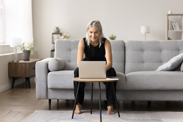 Obraz premium Smiling mature woman working from home sit at desk typing on laptop, solve issues remotely, correspond, e-mailing to client, engaged in workflow process. Modern wireless tech, internet usage, business