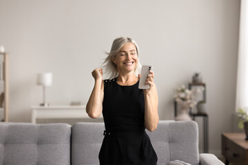 Cheery woman dancing in living room with smartphone in hand, user of digital streaming services...