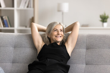 Smiling middle-aged female leaned on cozy sofa with hands behind hands, relish pleasant thoughts or...