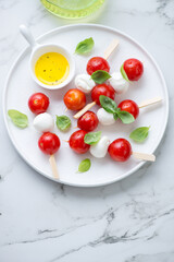 White plate with caprese skewers and olive oil, vertical shot on a white marble background, elevated view