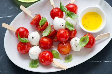 Caprese skewers with mozzarella balls, cherry tomatoes and green basil on a white plate, middle close-up, horizontal shot