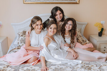 Four women are sitting on a bed, smiling and hugging each other. Scene is warm and friendly, as the women seem to be enjoying each other's company