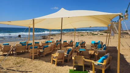 Vacation Seating Area by the Mediterranean Sea with Wooden Tables and Chairs, Blue Cushions, Sandy Beach, and Shade