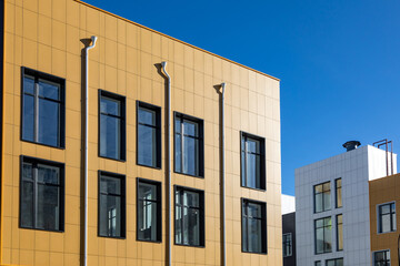 Drainpipes on the yellow ventilated facade. A complex of buildings of different storeys against a blue sky. Ventilated facades in different colours with a large glazing area