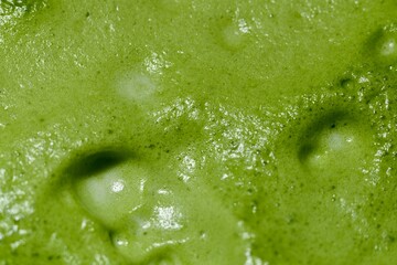 A close-up image of freshly made green tea, showing the vibrant green foam on its surface. The texture is smooth with visible bubbles, indicating the tea has been freshly whisked or blended.