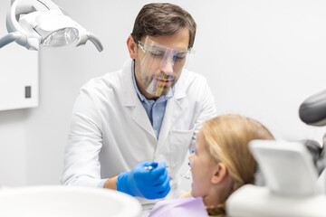 Obraz premium Little girl taking care of her teeth, sitting in chair while male dentist orthodontist in mask examining dental cavity