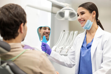 Positive man checking results of teeth whitening treatment in the dentist's office. Woman dentist holding mirror to satisfied male patient