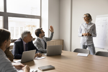 Group of businesspeople take part in educational seminar led by pretty mature female, male participant raising his hand, ask question or make comment, engaged in interactive, collaborative discussion