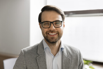 Head shot portrait happy millennial 30s businessman wear formal jacket and eyeglasses posing indoors, standing in office, smiling, looking at camera. Professional occupation person, career, promotion