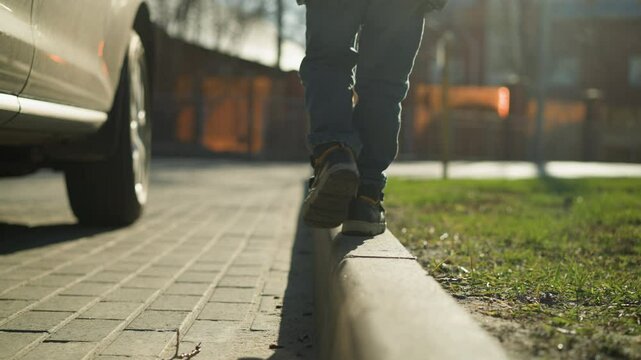A close-up of a child carefully balancing while walking along a pavement edge next to a car, dressed in a shiny black jacket, with blurred view of trees and buildings in the background