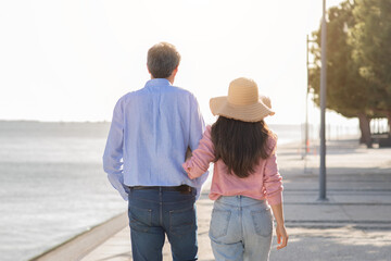 A couple walks hand in hand along a scenic waterfront promenade as the sun sets, creating a warm, romantic atmosphere. Their relaxed demeanor reflects a peaceful evening together.