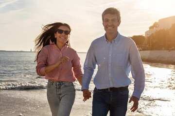 A joyful couple walks hand in hand along the shoreline, smiling and laughing as the sun sets, casting a warm golden glow over the ocean and creating a romantic atmosphere.