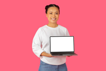 A cheerful young Asian woman smiles while holding a laptop with white blank screen against a vibrant pink backdrop.