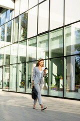 Business woman strolls confidently outdoors in smart attire, checking her messages against the backdrop of a modern glass building on a sunny day