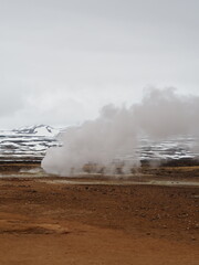 Visible steam from a geothermal fumarole contrasts against the stark, barren volcanic landscape with distant snow-covered hills and an overcast sky, creating a dramatic natural scene.