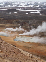 A rugged hilly geothermal field with steaming vents and patches of snow, highlighting the interplay of geothermal activity and the stark, rugged terrain.