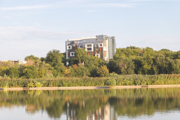 A modern apartment building with glass and metal facades stands on the bank of an industrial lake surrounded by trees and greenery