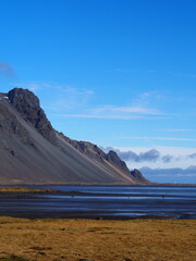 The image highlights a rugged, steep mountain with a flat grassy foreground, stretching out to a calm ocean in the distance under a vibrant blue sky.