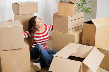 Young woman in her new house surrounded by cardboard boxes, they reach the ceiling. Happy, smiling and excited young girl opening moving boxes in student rental apartment.