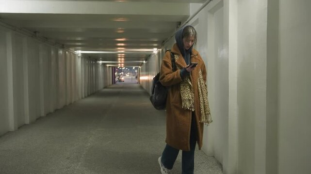 A girl in a brown coat and jeans walks through an underpass tunnel, leaning against the wall while looking at her phone with a worried and sad expression