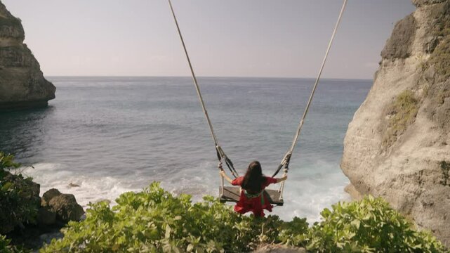Nusa penida island, Bali, indonesia. 4K video Young woman red dress Play on the swings at Diamond beach, beutiful diamond beach,Nusa Penida island. Popular travel destination on Bali, indonesia.