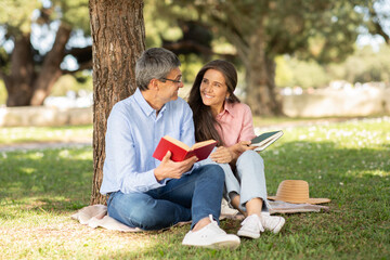 A couple enjoys a quiet afternoon under a tree, sharing their favorite books. The sun shines brightly as they smile and engage in conversation, surrounded by greenery.