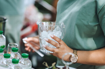 Young female bartender holds glass container with ice, tongs for making cold water, alcoholic cocktails. Photo, festive banquet.
