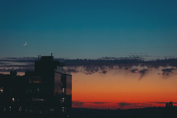 Obraz premium Cityscape with wonderful varicolored vivid dawn and the new moon. Amazing dramatic multicolored sky above dark silhouettes of city buildings at sunset. Atmospheric sunrise in overcast weather.