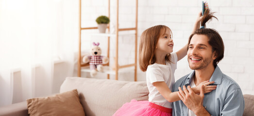 Fototapeta premium Pretty little daughter combing father hair, brushing and making horse tail, having fun together at home, copy space