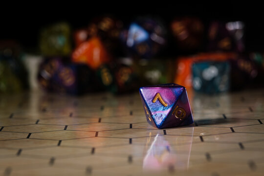 a blue and purple marbled eight-sided D8 dice with gold numbers, showing the number seven, sitting on a hexagonal tabletop gaming board with a variety of different types of dice in the background. 