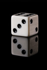 a white six-sided D6 dice with black dots, showing three dots, on a black background with a reflection