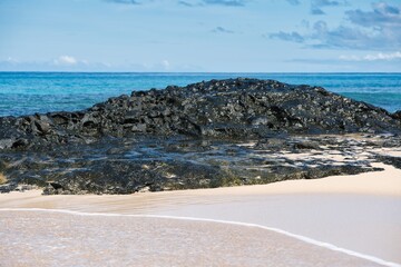 A huge dark and black rock made by volcanic activities on a sandy beach in Fiji with turquoise, blue water in the background. Black stones and rocks on a sandy beach. High contrast of stone and sand.