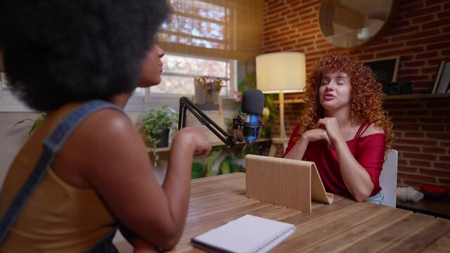 Multiracial women having a lively conversation while recording a podcast in a cozy studio with a microphone and tablet. Women Engaged in a Podcast Discussion in a Cozy Studio